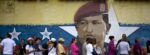 Mural on wall of Hugo Chavez's face, with a line of people standing in front of the wall
